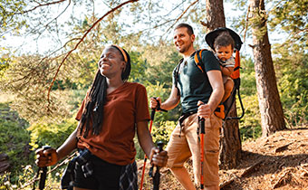 A couple and baby in a carrier smile while hiking in a sun-dappled forest