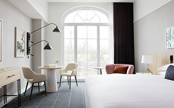 A light-filled hotel room with a king-size bed, a table with two chairs, and a large arched window